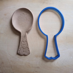 May include: A light brown clay spoon rest with a daisy design and bow accents, next to a blue spoon-shaped cookie cutter. The spoon rest is on a light brown wooden surface.