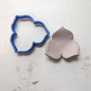 May include: A blue plastic cookie cutter in the shape of a three-petal flower sits next to a light purple clay flower. The clay flower is a three-dimensional version of the cookie cutter. The objects are on a white surface.