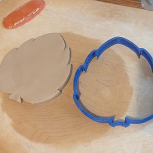 Puede incluir: Un cortador de galletas con forma de hoja de plástico azul junto a un trozo de arcilla con forma de hoja. Un trozo de arcilla naranja y oblonga está en la esquina superior izquierda. Los objetos están sobre una superficie de madera clara.