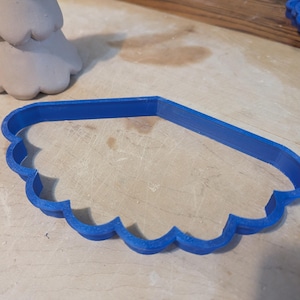 以下が含まれることがあります： 青いスカラップエッジのクッキーカッターが明るい表面にあります。部分的に見える、明るい灰色の粘土のオブジェクトが背景にあります。カッターの形状は雲または貝殻を連想させ、固体で不透明な材料で作られています。