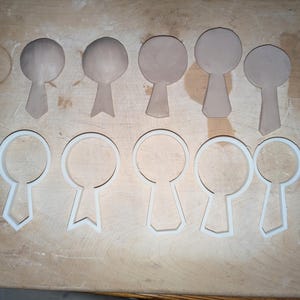 May include: A set of eight cookie cutters in the shape of a tie. The cutters are made of white plastic and are arranged in two rows on a wooden surface. The top row features five brown clay shapes that are similar to the cookie cutters.