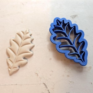 May include: A blue plastic leaf-shaped cookie cutter positioned next to a beige molded clay leaf cutout. The cutter features a detailed internal vein structure, while the clay piece displays the corresponding embossed leaf pattern.