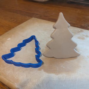 May include: A blue plastic Christmas tree cookie cutter next to a gray clay Christmas tree. The cookie cutter is on a wooden cutting board. The clay tree is a solid, three-dimensional shape.