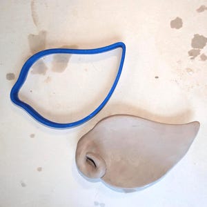 May include: A blue plastic cookie cutter in the shape of a leaf sits next to a light brown clay leaf. The clay leaf has a curled edge and a hole. The objects are on a light-colored surface.
