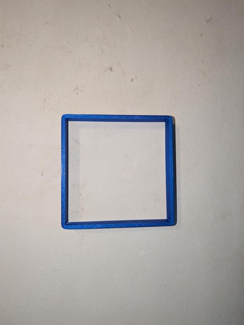 May include: A square, blue, 3D-printed object. The object is a hollow square frame, with a solid blue border. The frame is centered on a light gray surface.