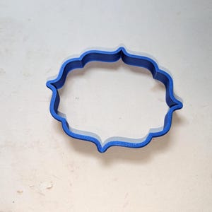 May include: A blue plastic cookie cutter featuring a decorative, symmetrical frame shape with curved edges and pointed corners. The object has a hollow center and a raised, thin wall structure designed for cutting shapes out of dough or clay.