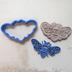 May include: Three bee-themed items: a blue cloud-shaped cookie cutter, a tan bee-shaped stamp, and a blue bee-shaped cookie cutter. The bee cutters have detailed wing and body patterns. The items are arranged on a light-colored surface.