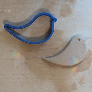 May include: A blue plastic bird-shaped cookie cutter next to a light-colored bird-shaped object. The objects are on a light-colored wooden surface. The bird-shaped object has a small hole near the top.