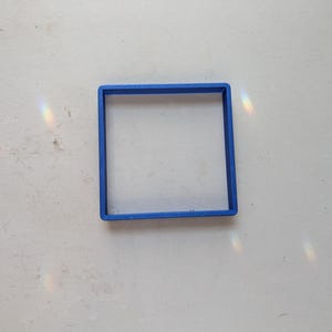 May include: A bright blue square-shaped cookie cutter. The cutter is made of plastic and sits on a white surface. The square shape is clearly defined, with clean lines and a uniform thickness. The cutter is designed for baking.