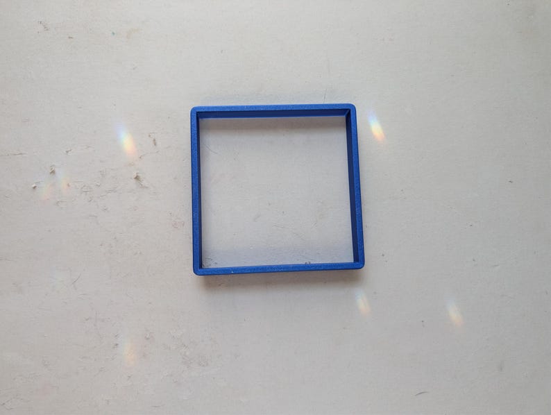 May include: A bright blue square-shaped cookie cutter. The cutter is made of plastic and sits on a white surface. The square shape is clearly defined, with clean lines and a uniform thickness. The cutter is designed for baking.