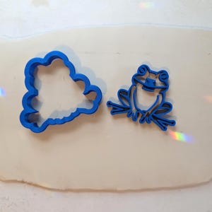 May include: Two blue plastic cookie cutters shaped like a frog. One cutter features a solid outline of a frog, while the other cutter depicts a detailed frog sitting on a branch. Both items are placed on a flat, light-colored, oval-shaped surface.