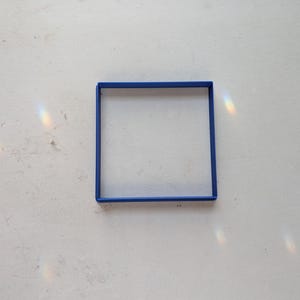 May include: A square, blue frame sits on a white, textured surface. The frame is a solid, vibrant blue, and the interior is empty. Light refracts, creating a rainbow effect on the surface.