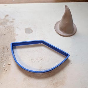 May include: A light brown clay witch hat and a blue plastic cookie cutter on a light-colored surface. The hat is conical with a wide brim. The cookie cutter is a curved shape with a flat side.
