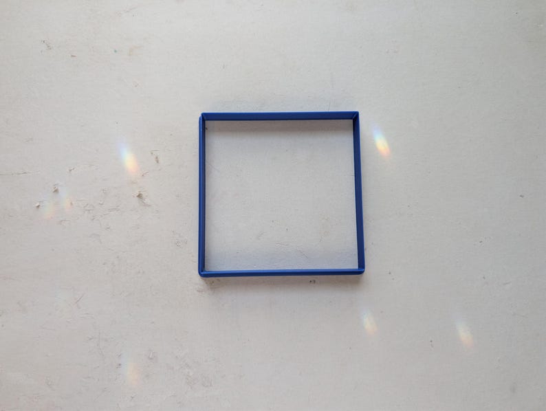 May include: A square, blue frame sits on a white, textured surface. The frame is a solid, vibrant blue, and the surface has subtle imperfections. Light refracts, creating a rainbow effect.