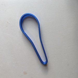 May include: A blue plastic cookie cutter in the shape of a spoon. The cutter is a solid, continuous line, with a rounded top and a tapered handle. The object is set against a white background.