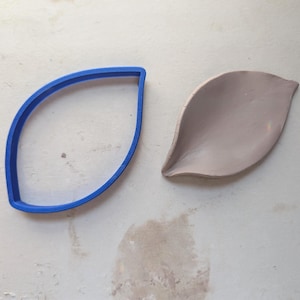 May include: A blue plastic leaf-shaped cookie cutter next to a gray clay leaf. The cutter is a solid blue outline of a leaf shape. The clay leaf is a light gray color with a curved shape.