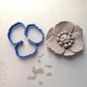 Puede incluir: Un cortador de galletas azul en forma de flor junto a una flor de arcilla clara y detallada. La flor de arcilla tiene múltiples pétalos y un centro texturizado. El cortador de galletas es un simple contorno de tres pétalos.