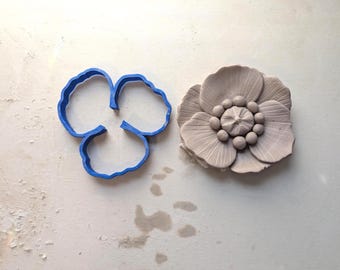 May include: A blue plastic flower-shaped cookie cutter next to a detailed, light-colored clay flower. The clay flower has multiple petals and a textured center. The cookie cutter is a simple outline of three petals.
