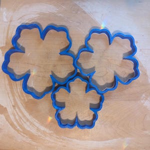 May include: Three blue flower-shaped cookie cutters of varying sizes are arranged on a light-colored wooden surface. The cutters have a simple, clean design, perfect for baking. The wooden surface has a few light-colored wooden planks on the left side.
