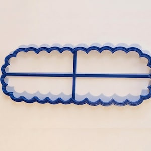 Op de afbeelding: Een blauwe plastic koekjesvorm in een langwerpige ovale vorm met een geschulpte rand. De vorm is voorzien van een centraal kruisvormig tussenschot dat de binnenkant in vier gelijke delen verdeelt voor een specifiek patroon in het deeg.