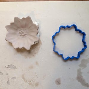 May include: A light-colored, flower-shaped cookie cutter next to a blue, flower-shaped cookie cutter. The flower-shaped cookie cutter is made of a light-colored material, and the other is made of blue plastic.