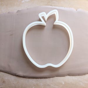 Könnte beinhalten: Weißer, plastikförmiger Ausstecher in Form eines Apfels auf einer braunen Oberfläche.