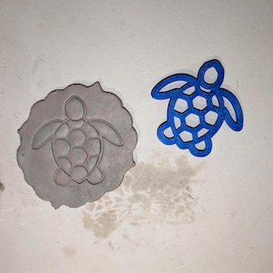 Puede incluir: Un sello de arcilla gris con forma de tortuga junto a un recorte azul con forma de tortuga. El sello tiene un diseño de tortuga prensado en la arcilla. El recorte azul tiene un patrón de panal en el caparazón de la tortuga.