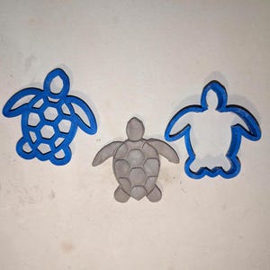 Puede incluir: Tres cortadores de galletas con forma de tortuga. Dos son de plástico azul, uno con un patrón de panal y el otro un contorno de espacio negativo. El tercero es un objeto gris con forma de tortuga.