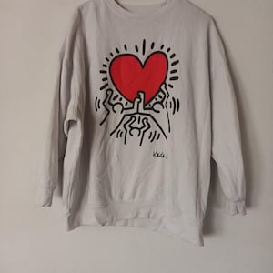 May include: A light gray sweatshirt featuring a large red heart design with black outlines and radiating lines. The design includes stylized figures holding up the heart. The artist's signature, "Keith," is visible.