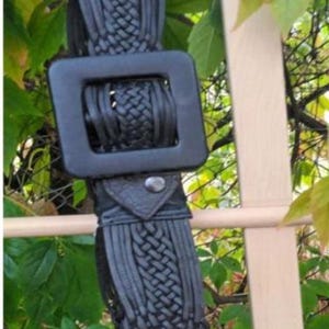 Puede incluir: Cintur&oacute;n negro tejido con una hebilla cuadrada grande. El cintur&oacute;n presenta un dise&ntilde;o trenzado y una correa de cuero con cierre de bot&oacute;n a presi&oacute;n plateado. El cintur&oacute;n cuelga sobre un fondo de hojas verdes y una estructura de madera.