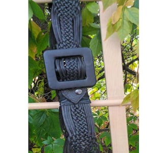 Puede incluir: Cintur&oacute;n negro trenzado con una hebilla cuadrada grande. El cintur&oacute;n est&aacute; hecho de cuero trenzado o material similar. La hebilla es de color negro mate. El cintur&oacute;n cuelga de una estructura de madera con follaje verde en el fondo.