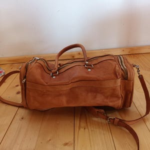Puede incluir: Una bolsa de lona de cuero marrón con un asa superior y una correa para el hombro desmontable. La bolsa tiene múltiples compartimentos con cremallera y un diseño clásico y vintage. La bolsa está hecha de cuero envejecido.