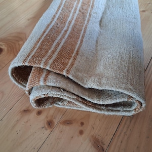 Op de afbeelding: Gevouwen beige en bruin gestreept textiel. De stof heeft een geweven textuur met verticale bruine strepen op een beige achtergrond. Het textiel is opgevouwen en toont de randen en lagen van de stof. Het textiel ligt op een houten oppervlak.