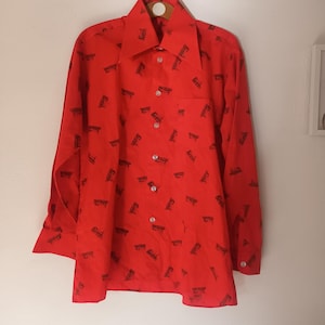 May include: A bright red button-down shirt with long sleeves and a pointed collar. The shirt features a pattern of small, dark brown shapes. The shirt has a chest pocket and is buttoned up. The shirt is hanging on a wooden hanger.