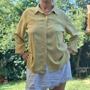 May include: A long-sleeved, button-down shirt in a muted mustard yellow color featuring a classic collar and a straight hemline. The shirt is paired with a white, lightweight skirt that has a delicate lace trim along the bottom edge.