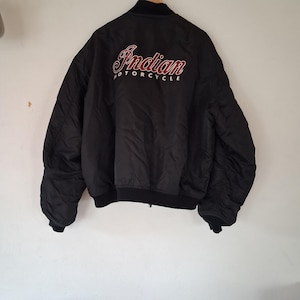 May include: A black bomber jacket featuring the text Indian Motorcycle embroidered on the back in red and white script and block lettering. The jacket has a ribbed black collar, cuffs, and hem, with a smooth, slightly gathered fabric texture.