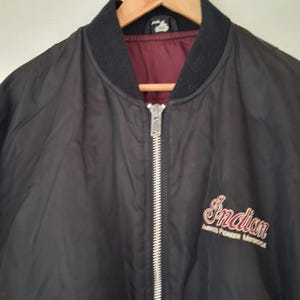 May include: A black bomber jacket with a ribbed black collar and a silver front zipper. The interior lining is a deep maroon color. The chest features embroidered text that reads Indian America's Pioneer Motorcycle in red and white lettering.