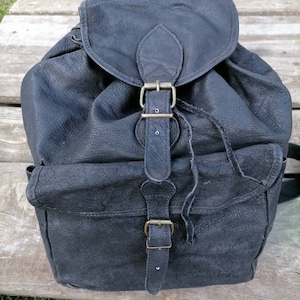 Puede incluir: Una mochila de cuero negro con cierre de solapa y detalles de hebilla. La mochila tiene un bolsillo frontal y bolsillos laterales. El cuero tiene un aspecto texturizado. La bolsa tiene un cierre de cordón y correas ajustables.