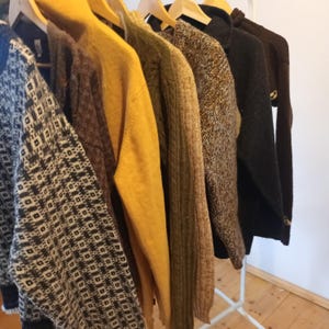 Op de afbeelding: Een kledingrek toont een verscheidenheid aan truien in bruine, gele en grijze tinten. De truien hangen aan houten kleerhangers en tonen verschillende texturen en patronen, waaronder een geometrisch ontwerp. Het rek is wit en de achtergrond is een effen muur.