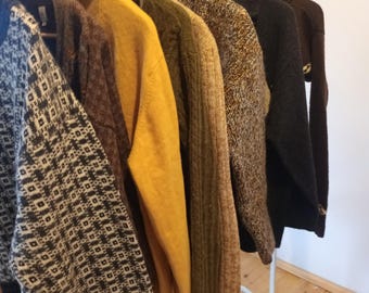May include: A clothing rack displays a variety of sweaters in shades of brown, yellow, and gray. The sweaters hang on wooden hangers, showcasing different textures and patterns, including a geometric design. The rack is white, and the background is a plain wall.