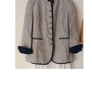 Op de afbeelding: Een grijze blazer met een verticaal streepjespatroon, met een opstaande kraag en donkerblauwe bies. De blazer heeft een rij knopen aan de voorkant en donkerblauwe manchetten.