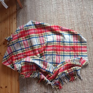 May include: A plaid blanket in red, blue, yellow, and white. The blanket has a fringed edge and is draped over a neutral-colored rug. The blanket is made of a soft material and is suitable for use as a throw or blanket.