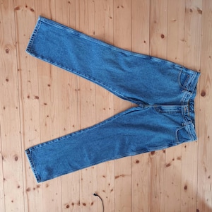 Pode incluir: Um par de calças de ganga azuis, colocadas numa superfície de madeira. As calças de ganga têm um design clássico de cinco bolsos com fecho de botão e fecho de correr. A ganga tem uma lavagem média, mostrando um desbotamento subtil.
