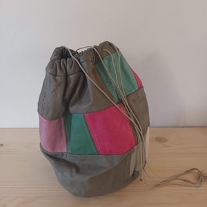 Op de afbeelding: Een patchwork leren trekkoordtas in tinten grijs, roze, groen en beige. De tas heeft een afgeronde vorm en is aan de bovenkant samengeknepen met een trekkoordsluiting. De tas is gemaakt van verschillende aan elkaar genaaide leren lappen.