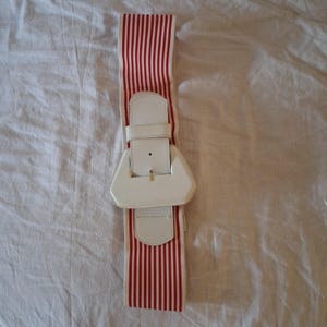 May include: A wide elastic belt featuring a pattern of vertical red and white stripes. The belt includes a large, white, geometric-shaped buckle with a central strap and a single prong, designed to secure the band around the waist or hips.