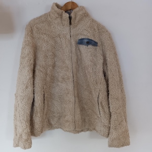 May include: A beige, zip-up fleece jacket with a high collar and a small blue plaid pocket on the chest. The jacket is hanging on a wooden hanger against a white background. The fleece material appears soft and textured.
