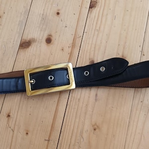 May include: A black leather belt with a gold-colored rectangular buckle. The belt has a classic design with a simple, elegant aesthetic. The belt is approximately 1.5 inches wide.