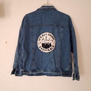 Puede incluir: Una chaqueta de mezclilla azul con un parche circular grande en la parte posterior. El parche es blanco con bordado negro que representa un horizonte urbano, estrellas y el texto "IT'S ALL GOOD KEEP PEACE" en un círculo alrededor.