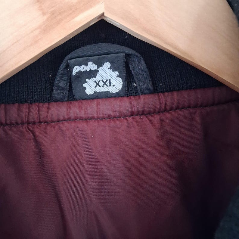 May include: A black jacket interior featuring a dark maroon lining and a black fabric neck label. The label displays the text "polo" in a white stylized font above the size "XXL" in white capital letters. The jacket is hanging on a wooden hanger.