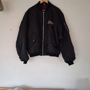 May include: A black bomber jacket featuring a silver front zipper, ribbed cuffs and hem, and two front pockets with snap closures. The chest displays white and red embroidered text that reads Indian, with a utility pocket on the left sleeve.